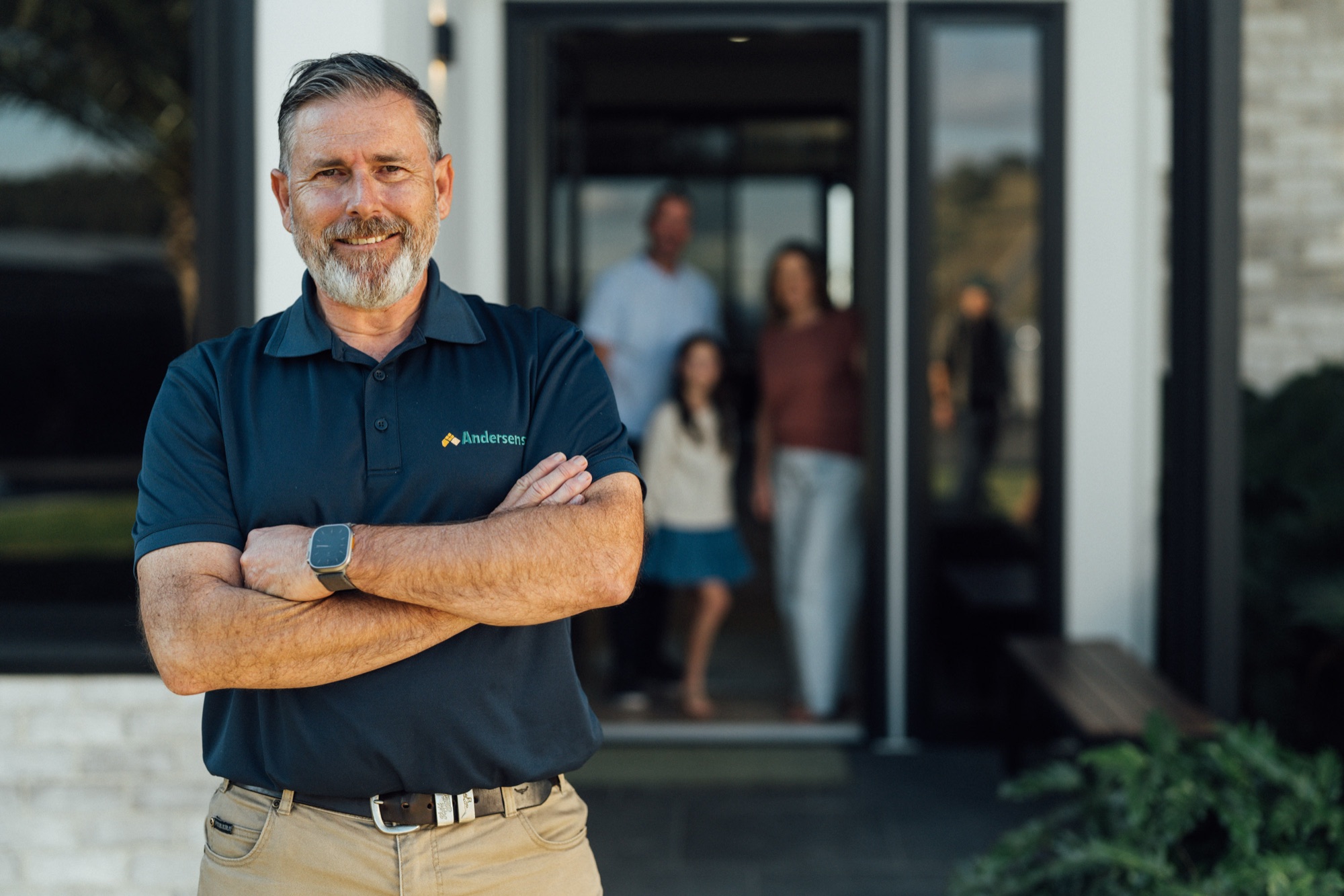 An Andersens consultant standing with arms folded at the entrance of a home, with a family behind
