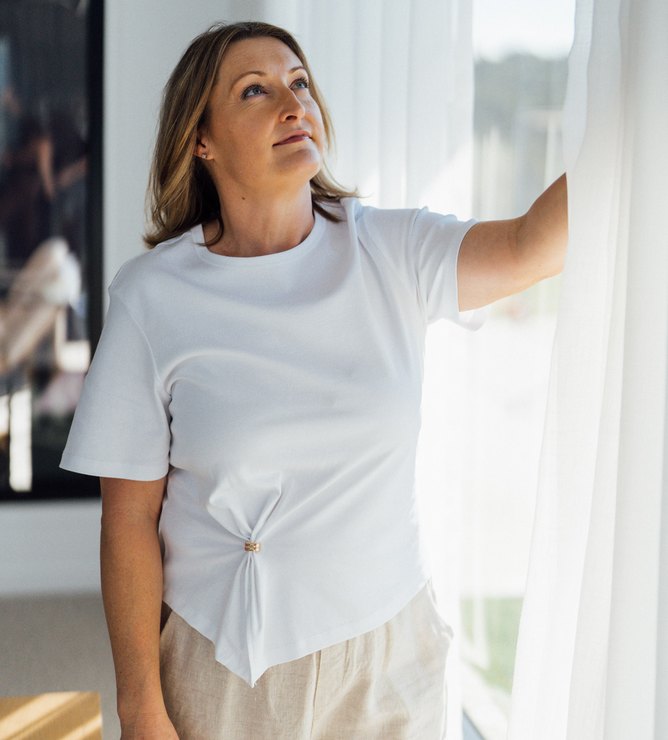 A woman drawing a sheer curtain back from a sunlit window