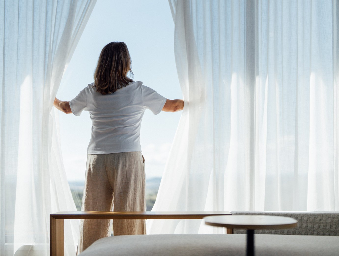 A woman holding sheer curtains open to a view over the countryside