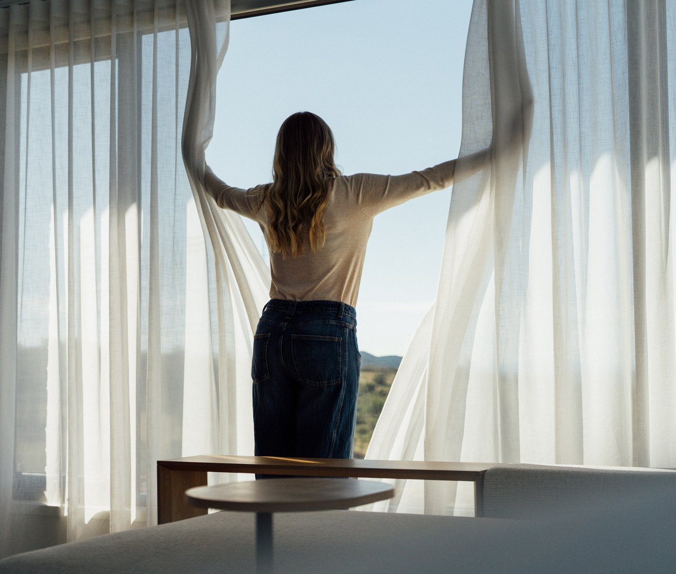 A woman drawing sheer curtains open across a full-height window onto a rural view