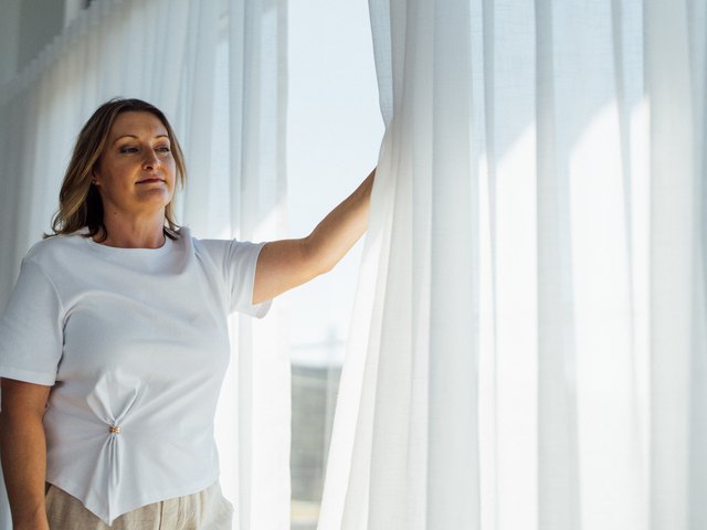 A woman standing at a window with sheer curtains in daylight
