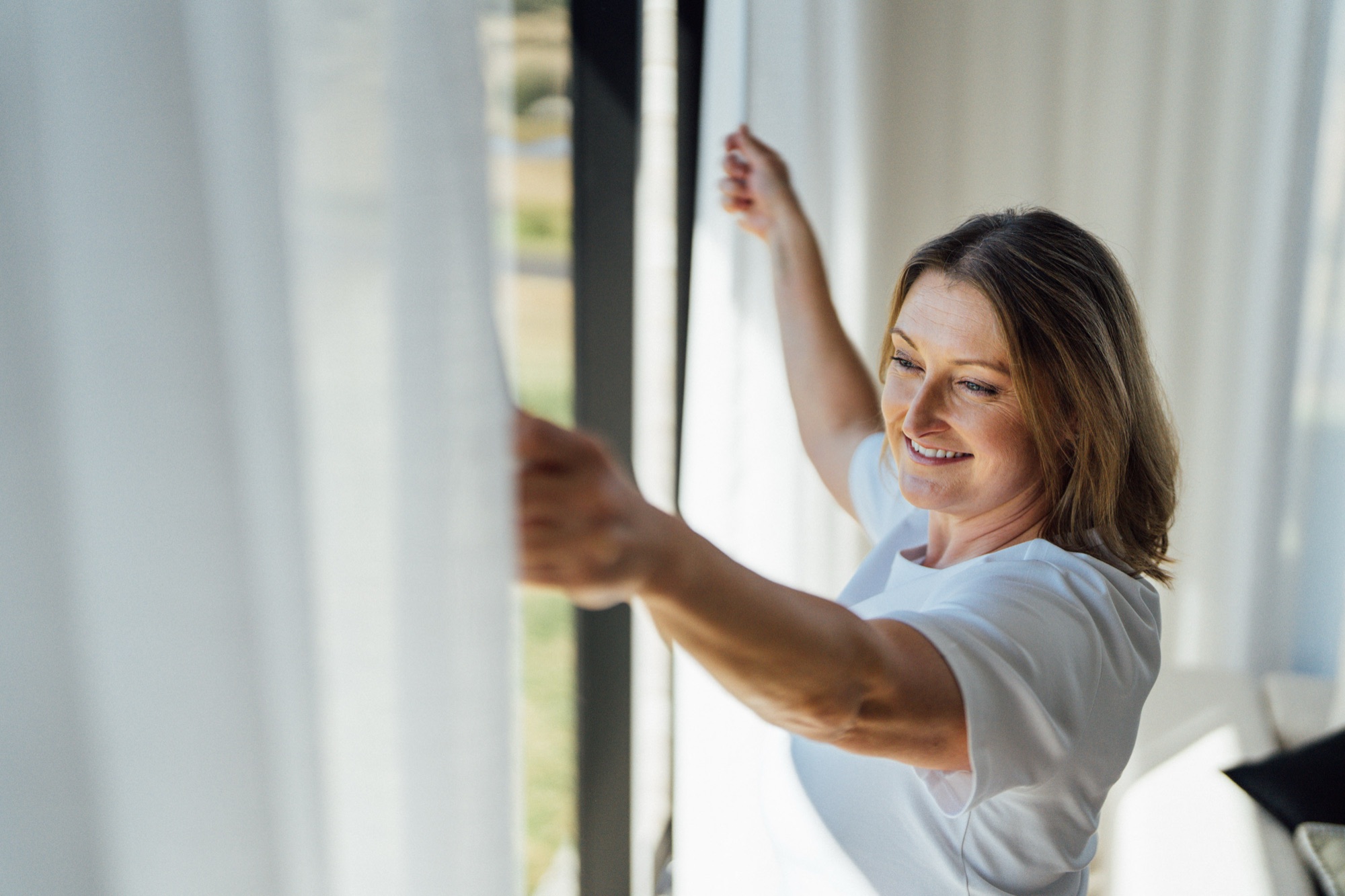 A woman drawing a sheer curtain across a window in her living room