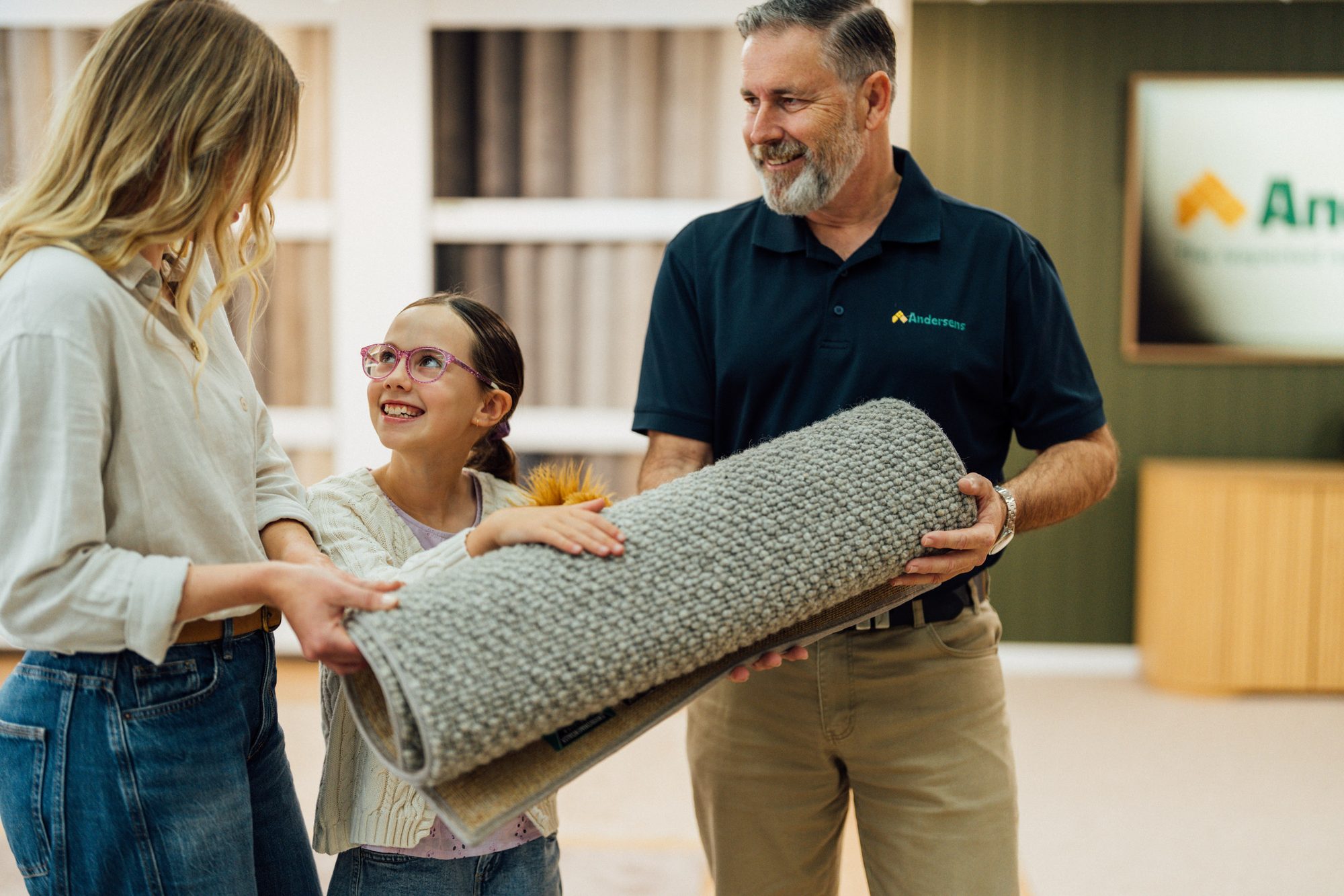 An Andersens team member carrying a roll of carpet with a mother and daughter in the showroom