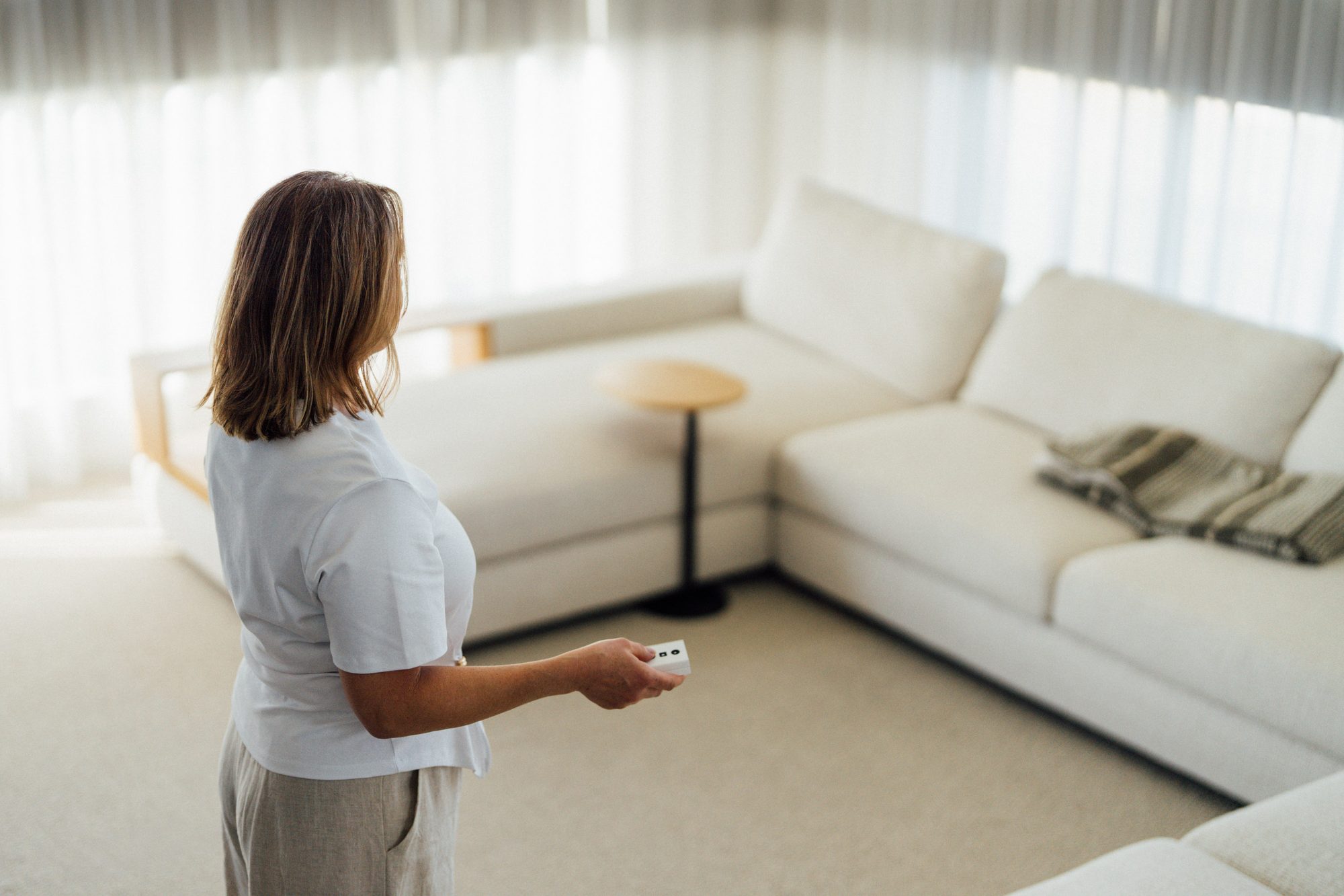 A woman using a remote to adjust the window furnishings along one wall of a carpeted living room