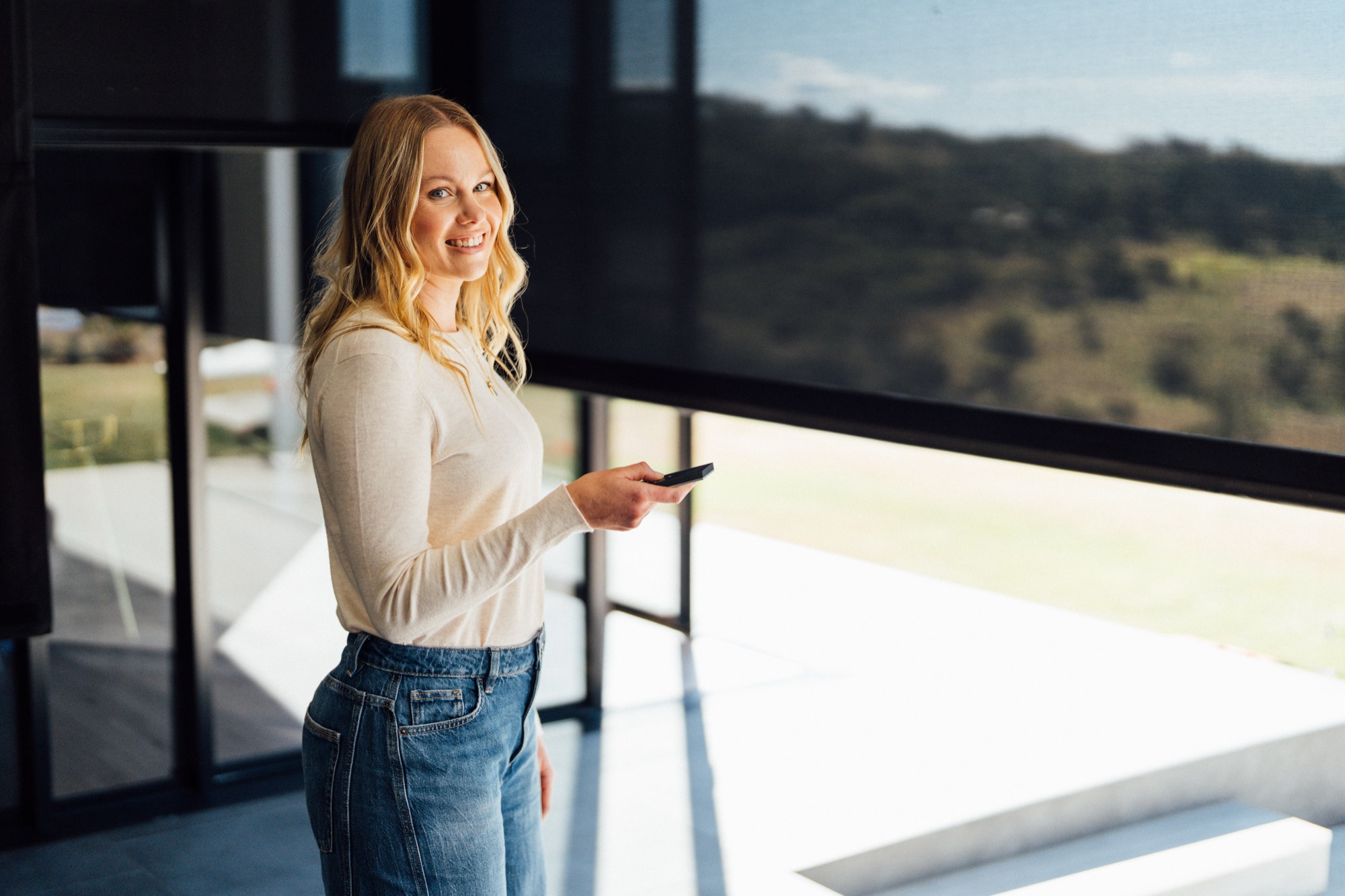 A woman holding a remote control beside full-height windows in a living room