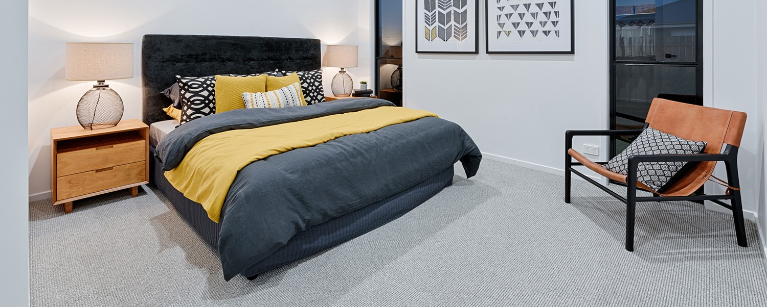 A carpeted master bedroom with a dark bedhead, bedside lamps and a leather chair