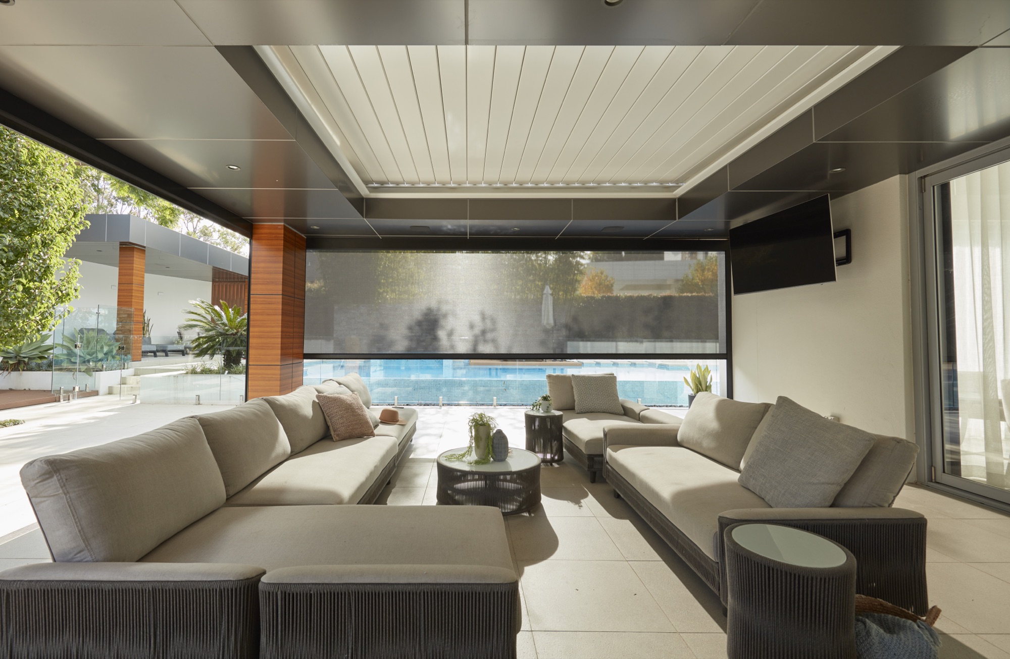 An outdoor lounge under a covered terrace, with an exterior screen lowered across the opening towards a pool