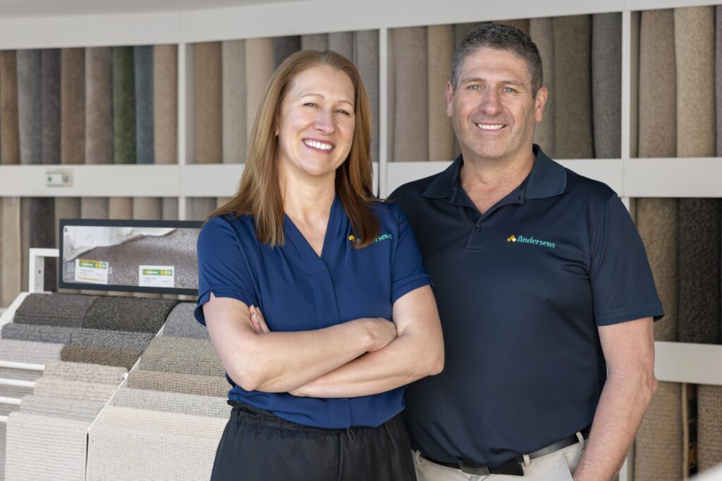 The Andersens Fyshwick owners smiling in front of the carpet wall