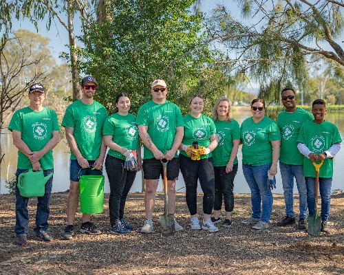 A group of ten volunteers in green shirts standing with spades and a watering can beside a lake