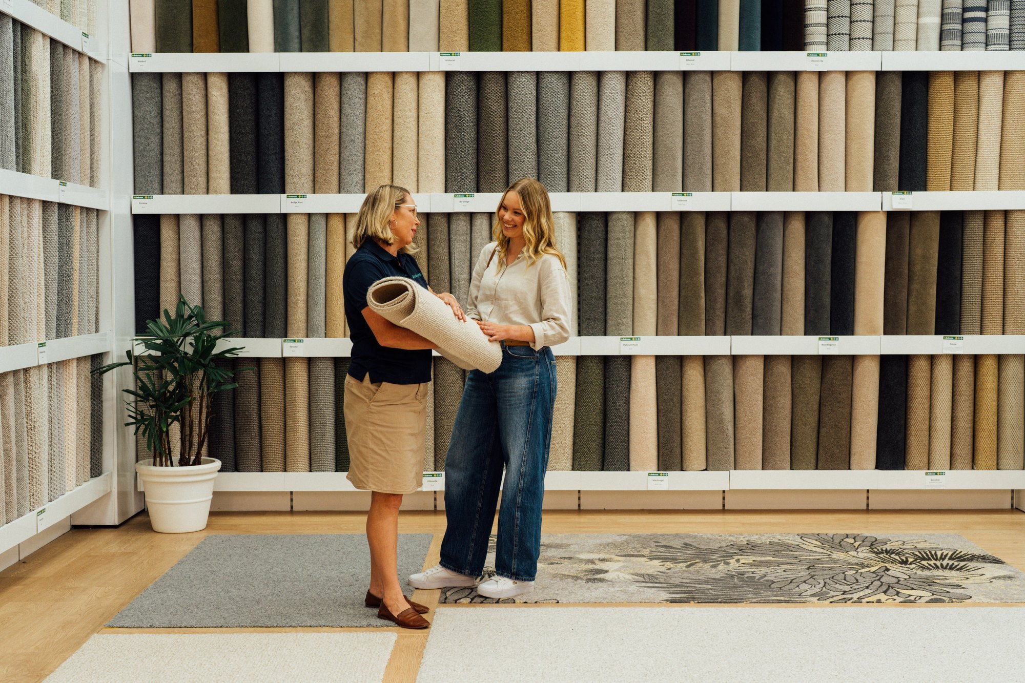 Two people beside floor to ceiling walls of carpet samples in an Andersens showroom, with rugs laid out on the floor